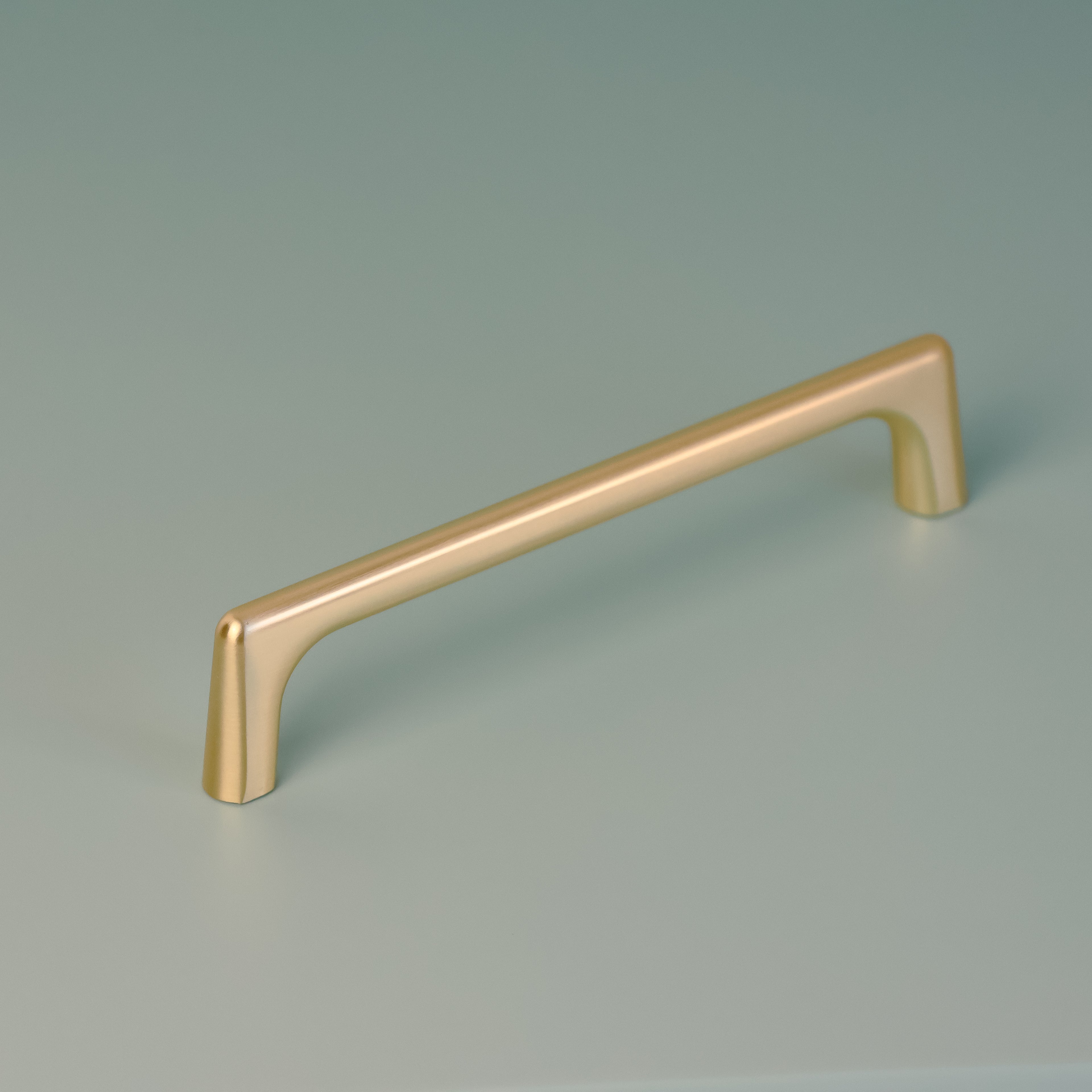 Darwin 128mm cabinet handle in brushed brass finish, displayed on a soft green background, showcasing its sleek, minimalist design and gently curved edges.